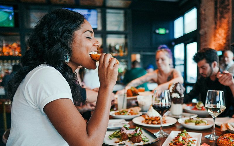 A woman out to eat with her friends, with many plates of tapas and glasses of wine between them. She is excitedly biting into one of the tapas, maybe a crostini.