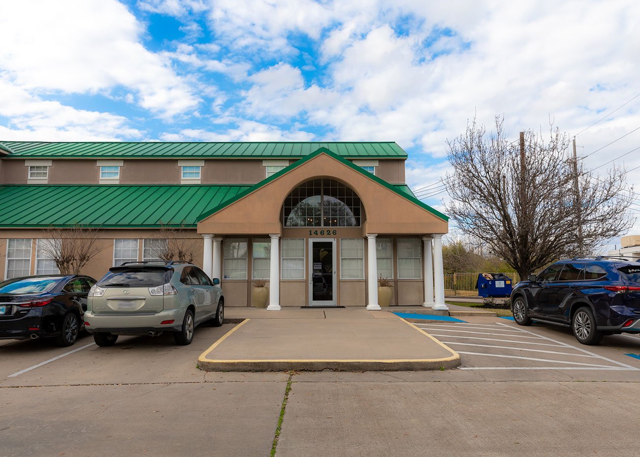A beige two-story building with a green metal roof and a covered entrance supported by white columns. The building number 14626 is above the doorway. There are several parked cars in front, including a black sedan, a gray SUV, and a blue SUV. The sky